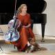Cellist Harriet Krijgh sits with her cello and a dog in front of a grand piano on a concert stage.