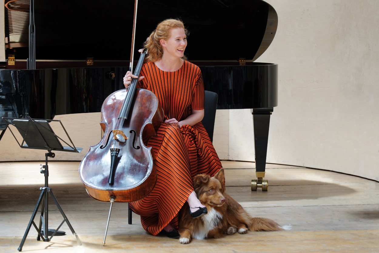 Harriet26©Armin Dreiling Cellist Harriet Krijgh sits with her cello and a dog in front of a grand piano on a concert stage.